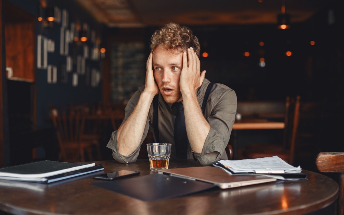 A stressed man with hands on head, surrounded by work materials, sits in a dimly lit restaurant.