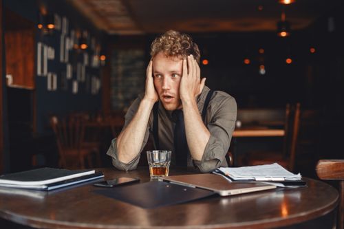 Free A stressed man with hands on head, surrounded by work materials, sits in a dimly lit restaurant. Stock Photo