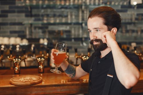 Free A bearded man sipping craft beer at a stylish pub with a lively bar counter. Stock Photo