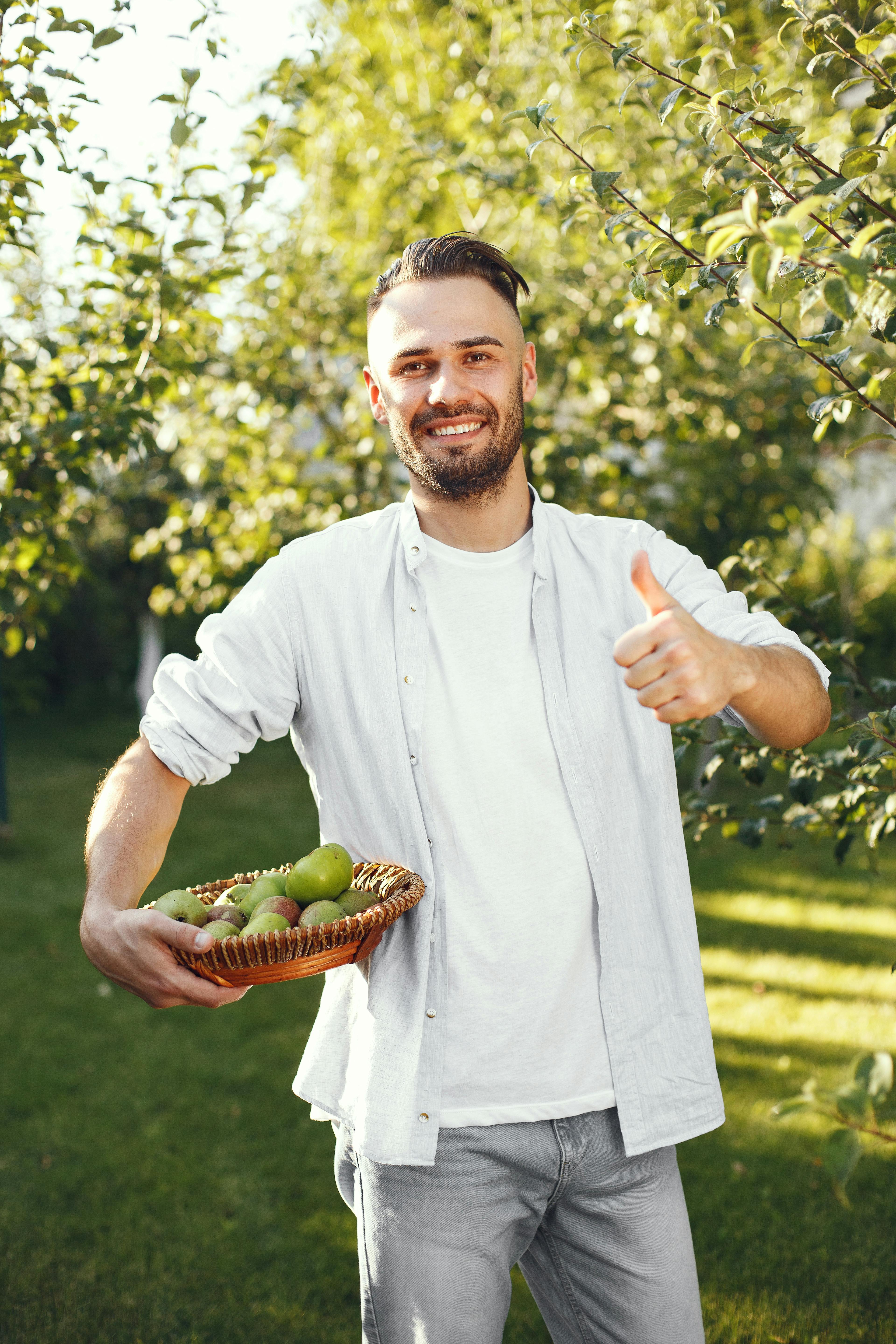 Happy man holding a basket of apples in a sunny garden, giving a thumbs up.