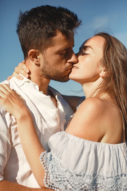 Free A young couple shares a tender kiss outdoors under a bright blue sky, exuding warmth and love. Stock Photo