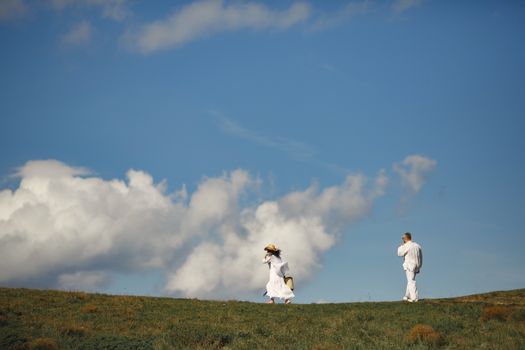 A couple dressed in white enjoys a sunny walk in a grassy field under a blue sky.