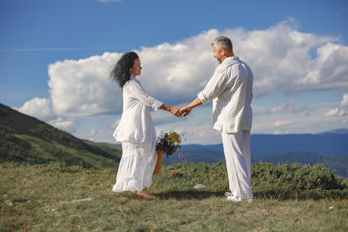 Free Man in White Dress Shirt and Woman in White Dress Holding Hands on Green Grass Field Stock Photo