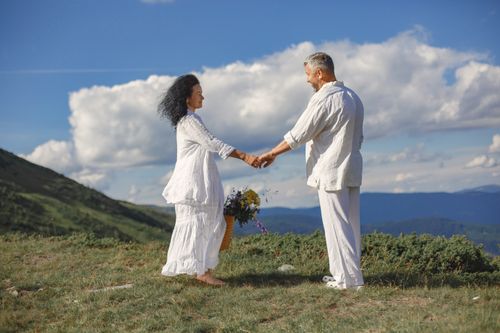 Free Happy couple in white attire holding hands outdoors in a scenic mountain landscape. Stock Photo