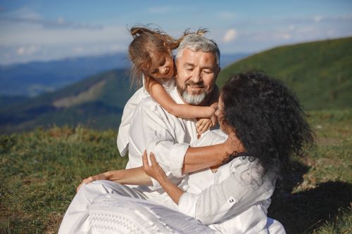 Free A joyful family moment outdoors, showing grandparents hugging their granddaughter under a sunny summer sky. Stock Photo