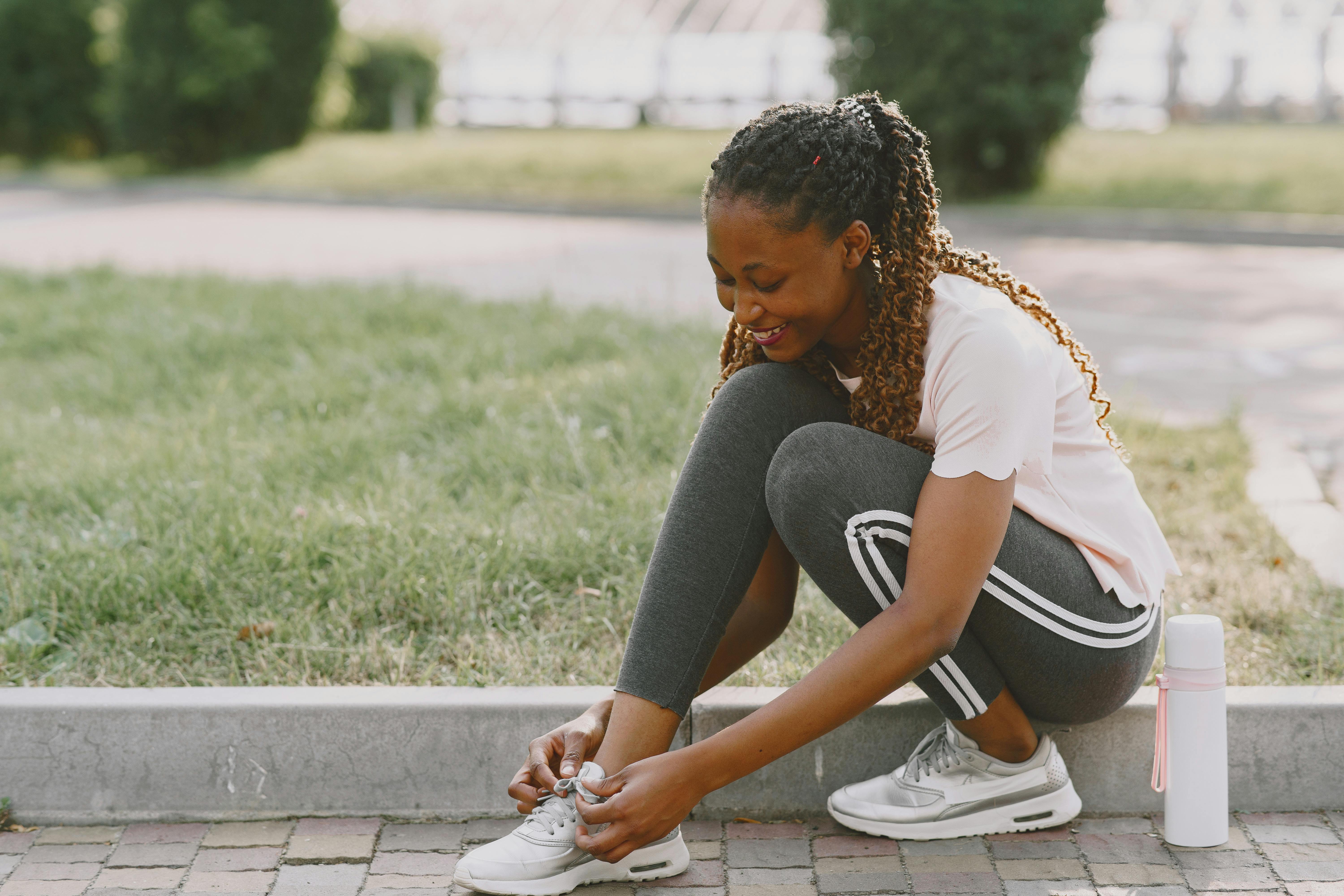 Young woman smiling while tying running shoes, bright park background, sunny day, fitness journey start.