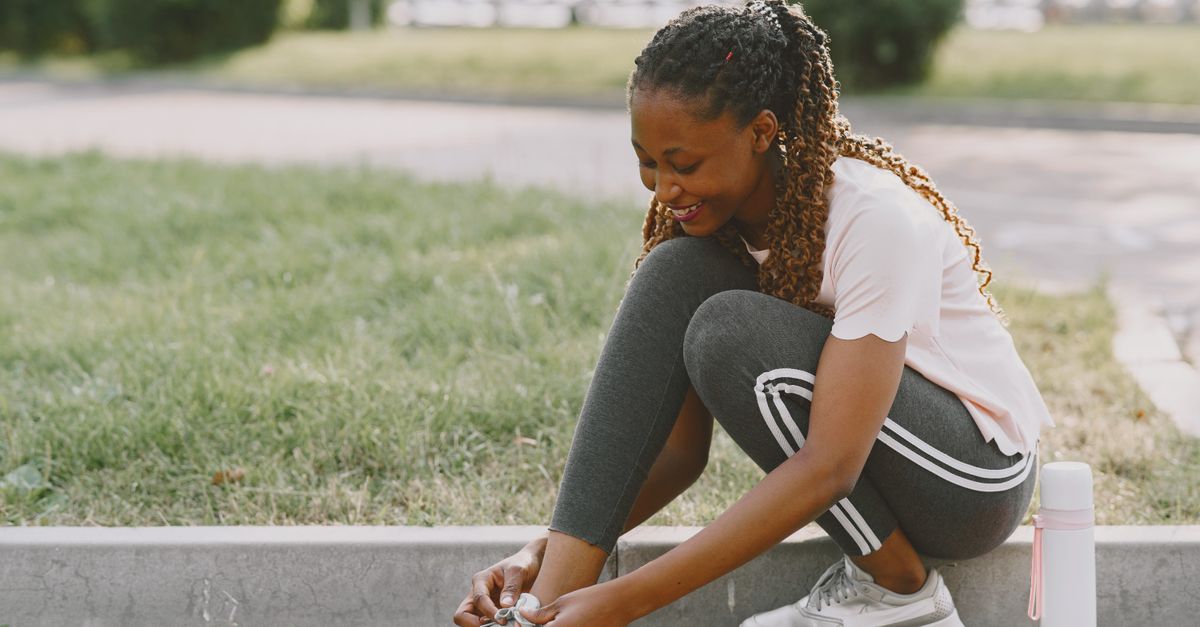 Smiling woman tying her sneakers while sitting outdoors on a sunny day, ready for a workout.