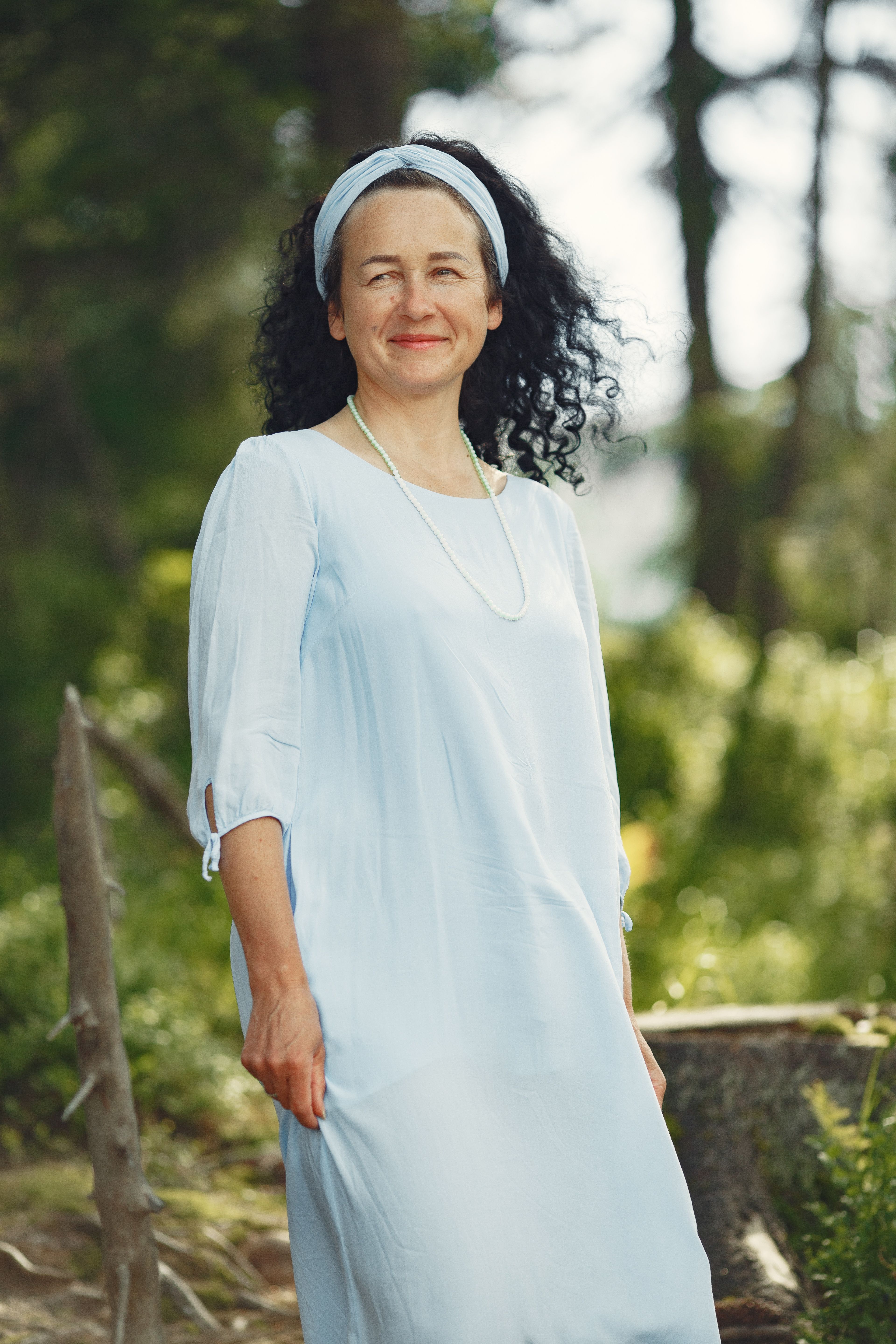 Adult woman in a white dress with a headband standing in a natural setting.