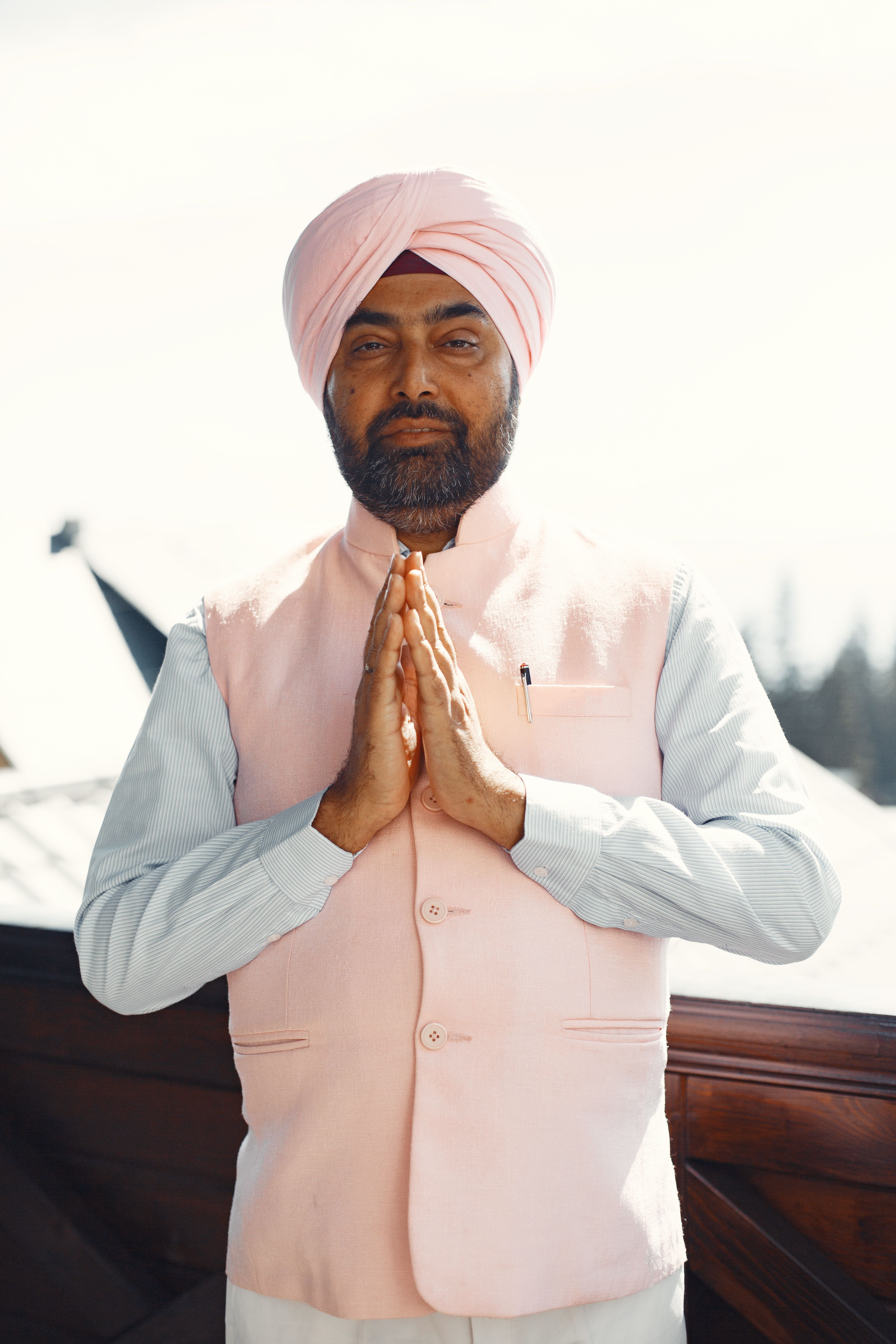 A man wearing a pink turban and vest stands outdoors with praying hands.