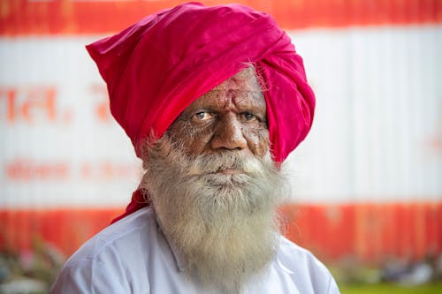 Elderly ethnic bearded male in bright national red turban looking at camera on blurred background
