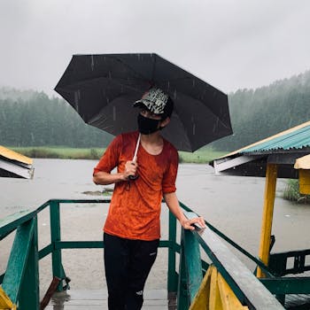 Man standing on a boardwalk under an umbrella during rainy weather by a lake.
