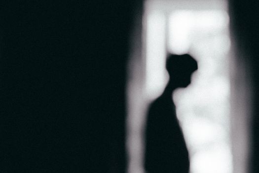 Moody black and white silhouette of a person standing by a window, conveying solitude.