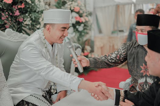 Groom in traditional attire shaking hands during wedding ceremony with floral decor.