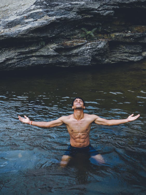 Shirtless ethnic male enjoying nature while sitting in river against rocky shore at daytime