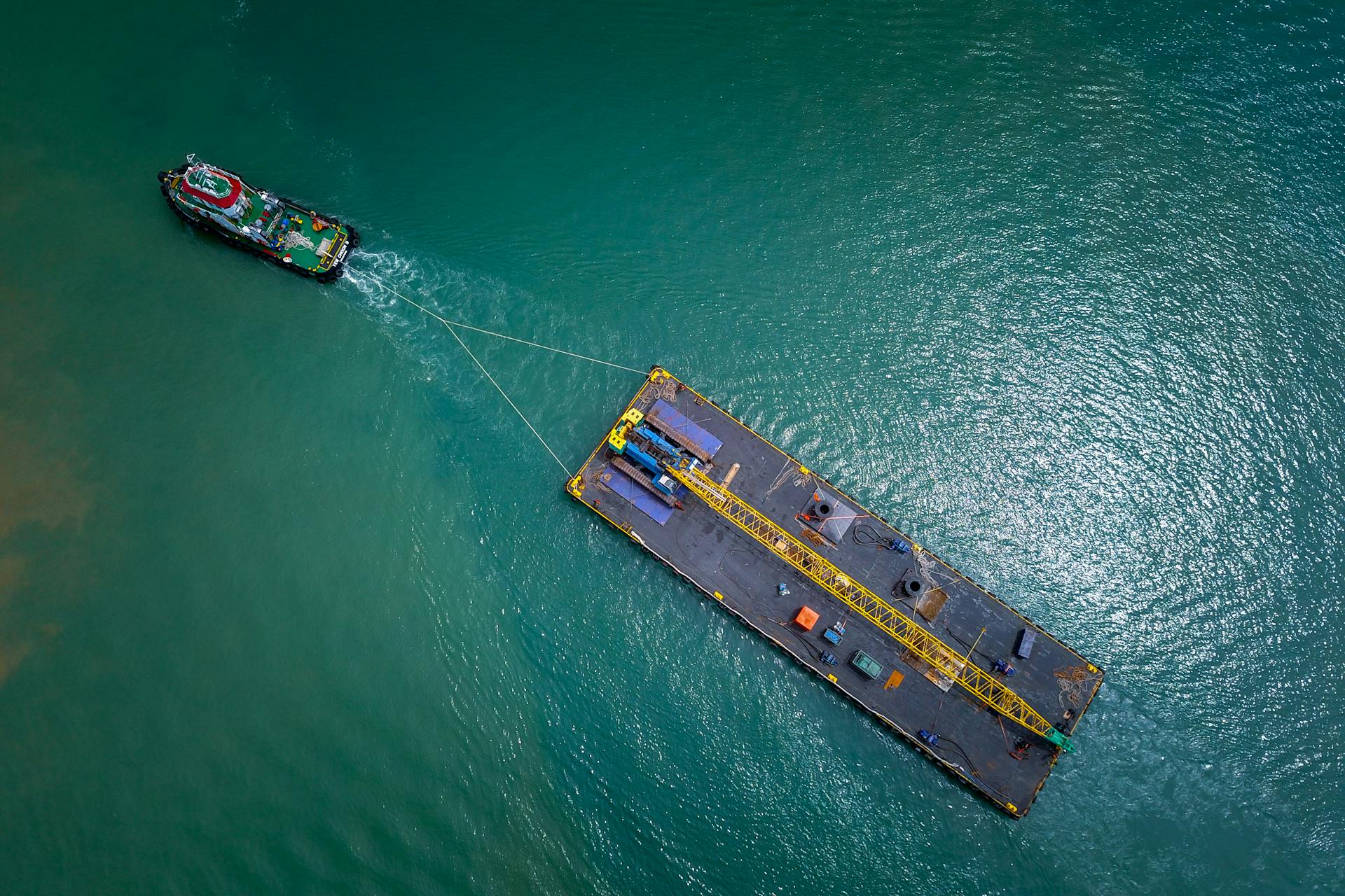 Tugboat towing a barge across turquoise ocean waters, captured from above.
