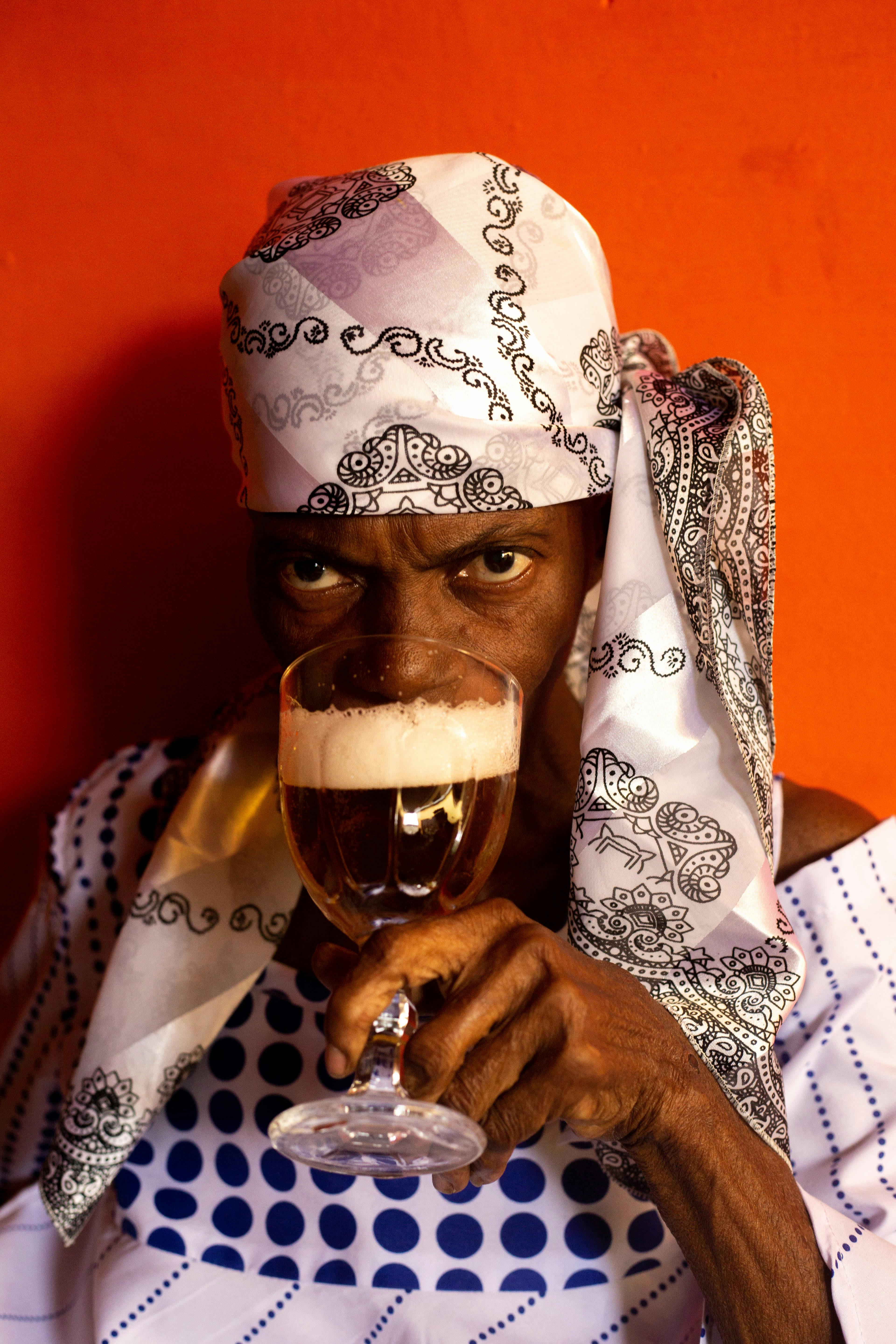 Concentrated crop ethnic female in headscarf drinking foamy alcohol beverage from glass covering half face and looking at camera while standing on red background
