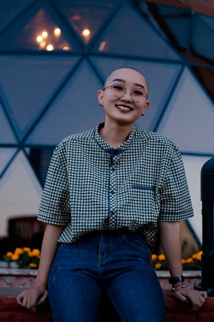 Happy young bald headed androgynous person in shirt and jeans looking at camera with bright smile while leaning on curb next to flowerbed and modern futuristic construction