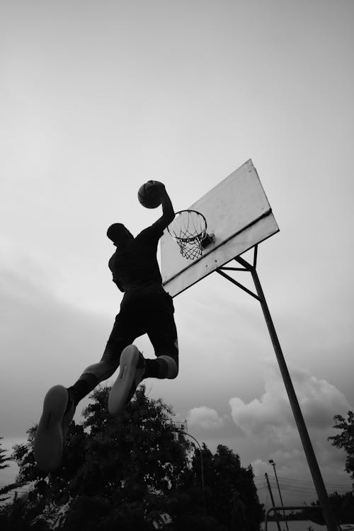Black and white full body of tall basketball player jumping and throwing ball in hoop against gray sky with clouds