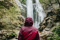 白天，一位穿着红色外套、戴着兜帽的匿名游客站在岩石悬崖旁的水流附近，这是他的后视图