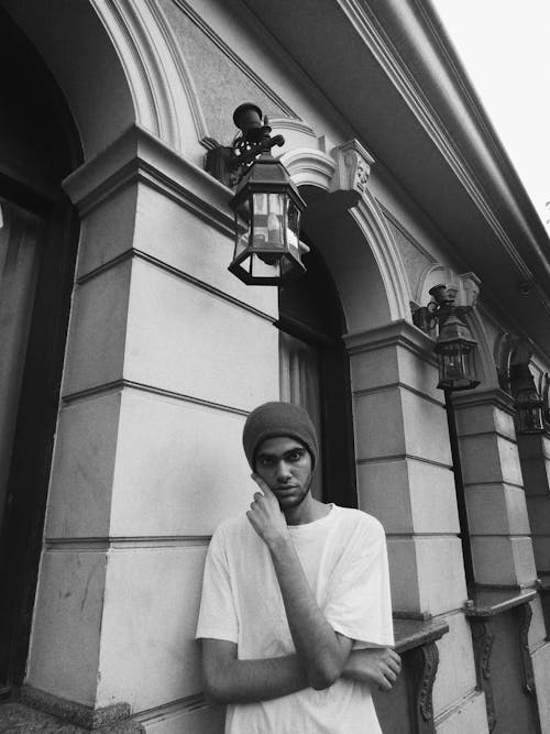 Black and white serious confident ethnic male in street style clothes touching face and looking at camera while standing outside old arched building