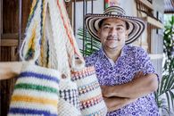 A smiling man with a traditional hat poses near colorful bags in a Colombian market.
