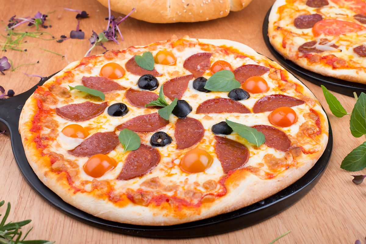 Salami pizza with black olive slices, halved cherry tomatoes and basil on a dark pan