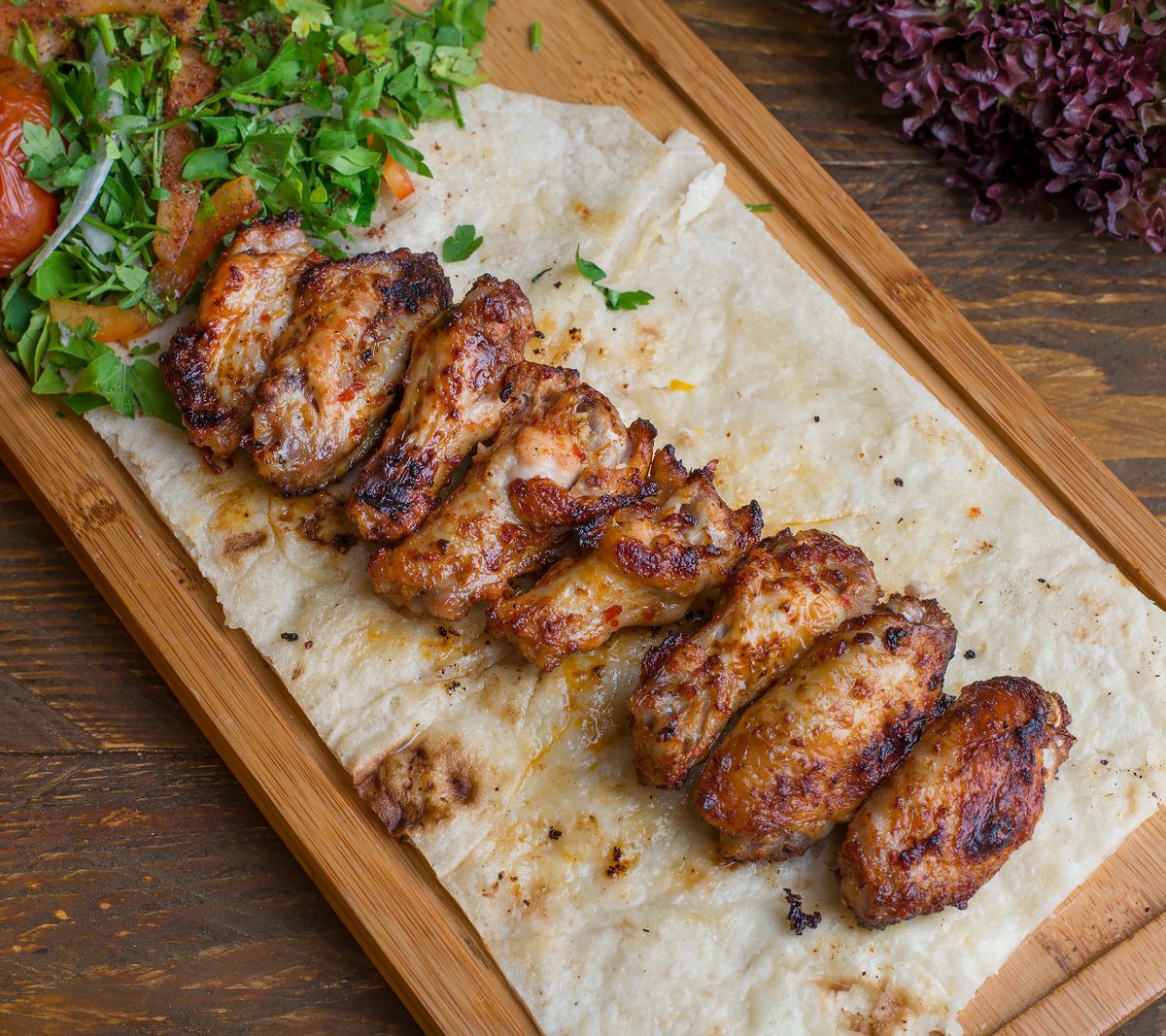 Grilled chicken laid over charred flatbread on a wooden board with herbs and salad. Photo by Shameel mukkath on Pexels.