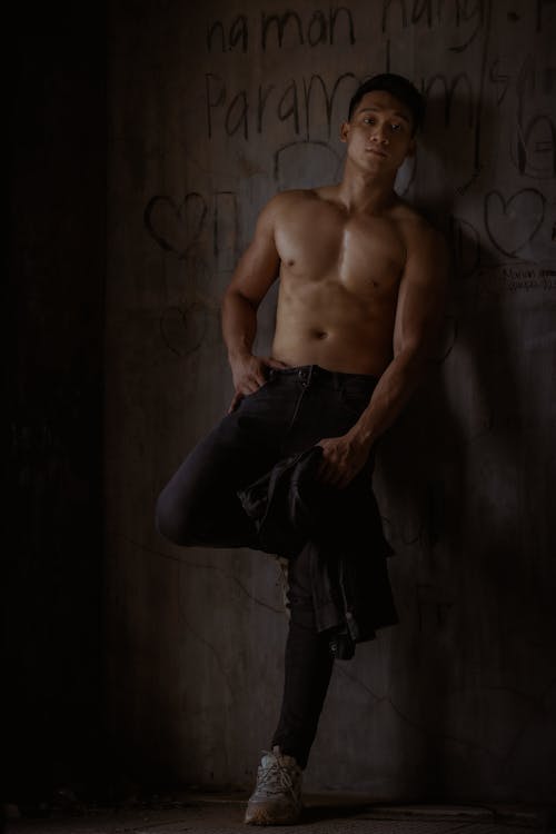 Full body of shirtless male in sporty pants looking at camera while leaning on concrete wall with drawing in abandoned building with hand on hip and leg bent