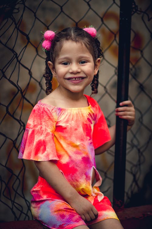 Adorable kid wearing vivid dress sitting against mesh fence and smiling while looking at camera