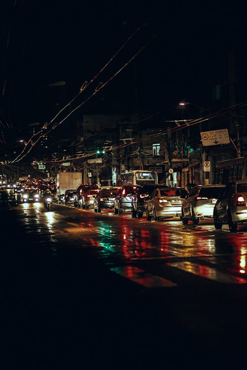 Modern cars with glowing lights in traffic jam on asphalt road on street with buildings and trolley wires at night
