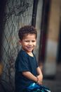 Adorable child with curly hair in casual clothes smiling while looking at camera against mesh fence at daytime