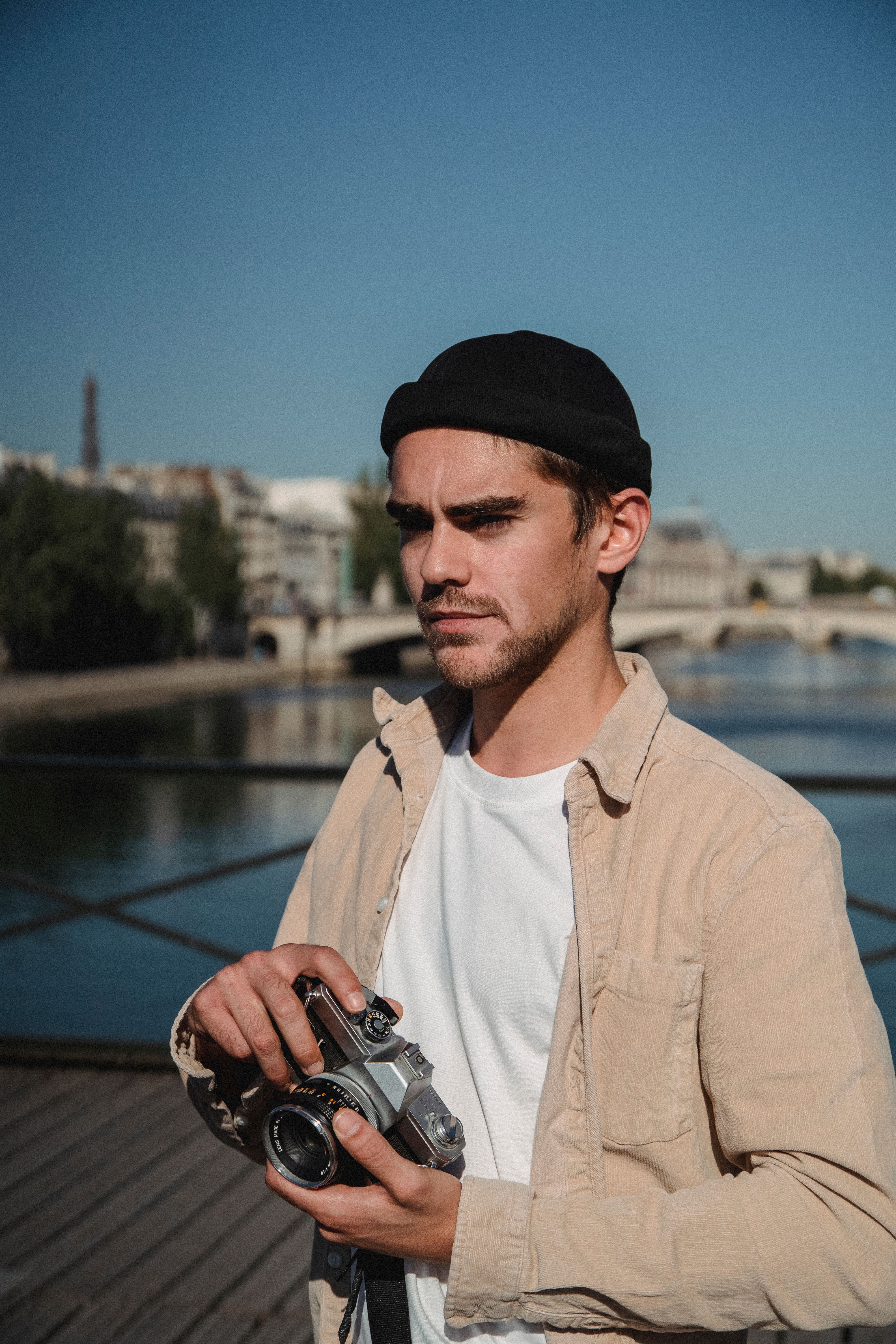Man in casual attire holding a vintage camera on a bridge with cityscape in background.