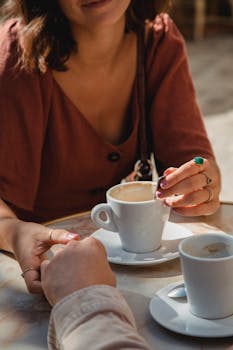 Two people enjoying coffee at a café, holding hands over mugs in a relaxed atmosphere.
