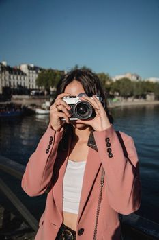 Stylish woman in pink blazer taking photos by the riverside on a sunny day, capturing the picturesque scenery.