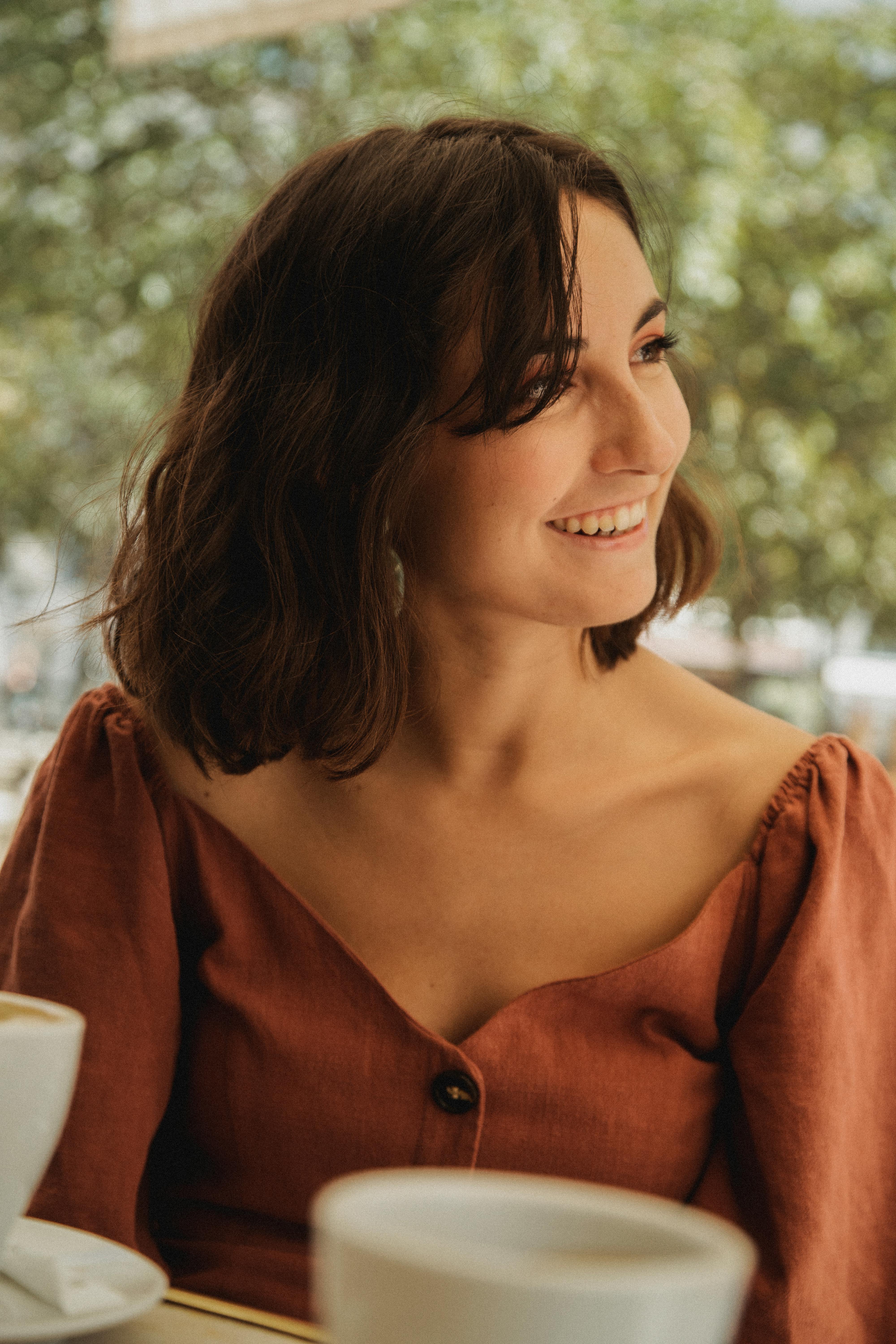 A woman enjoying coffee outdoors with a smile, surrounded by greenery and sunlight.