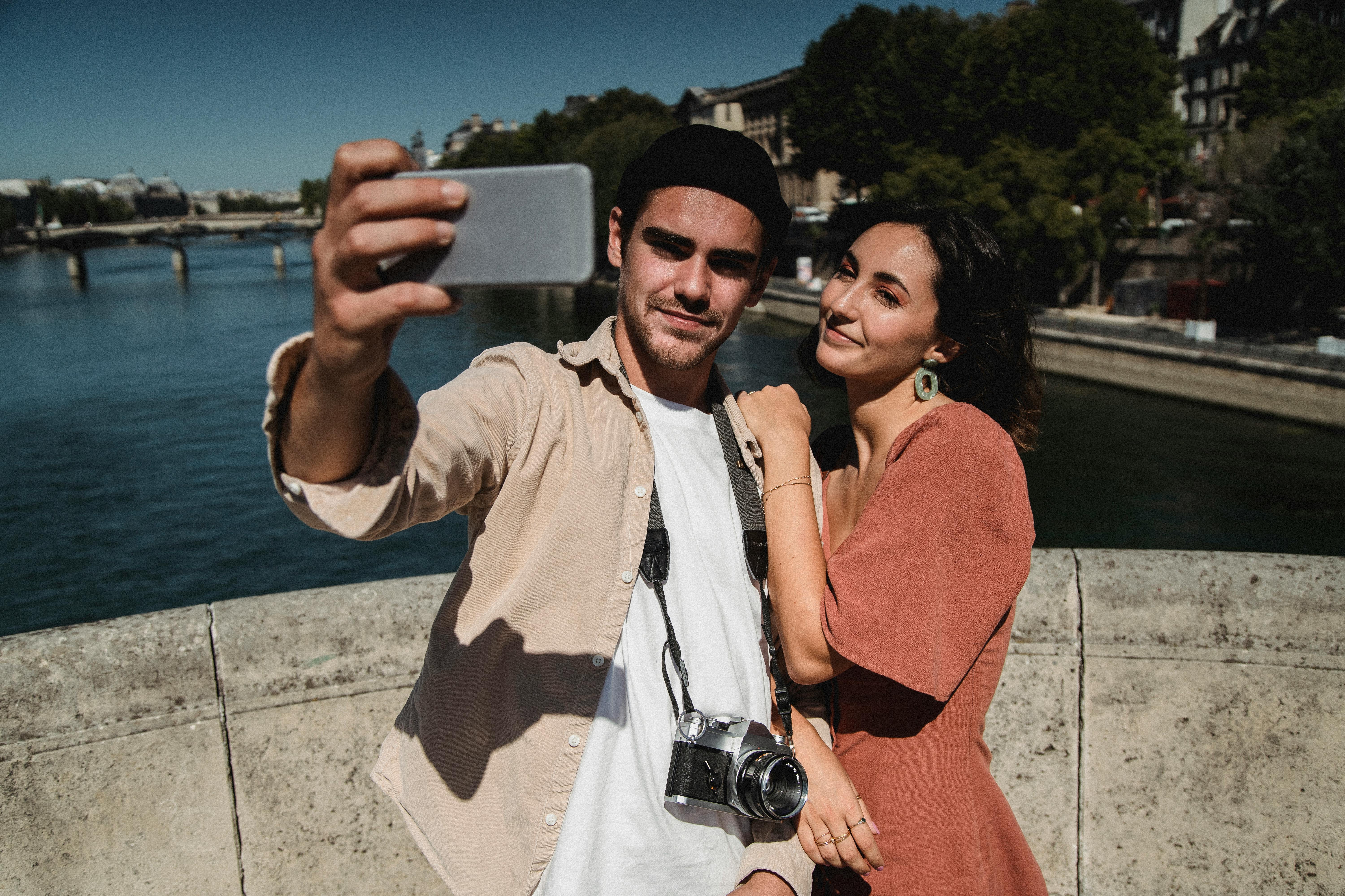 A happy couple takes a selfie on a scenic riverside bridge, enjoying a sunny day.
