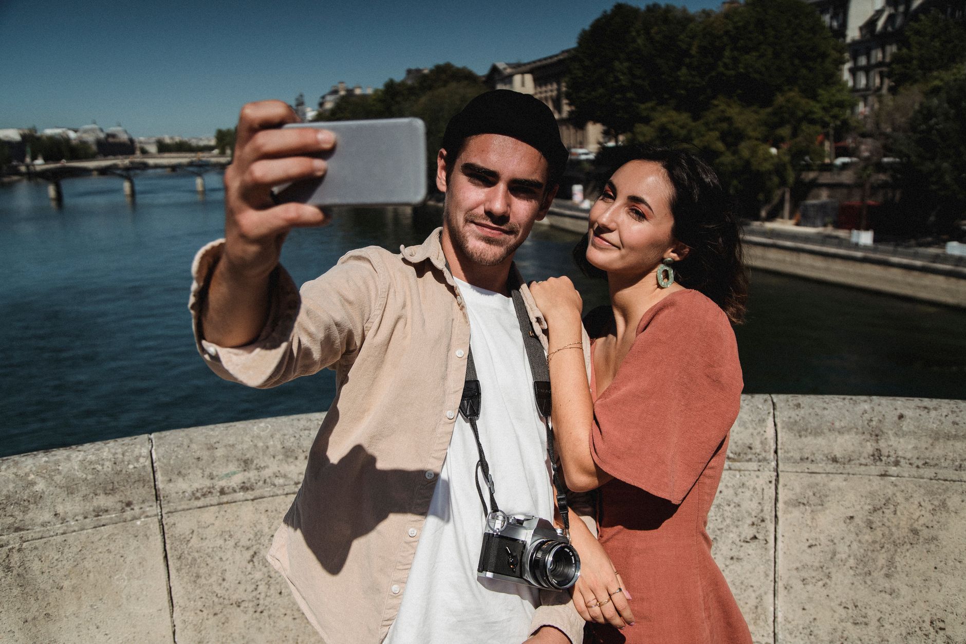 A happy couple takes a selfie on a scenic riverside bridge, enjoying a sunny day.