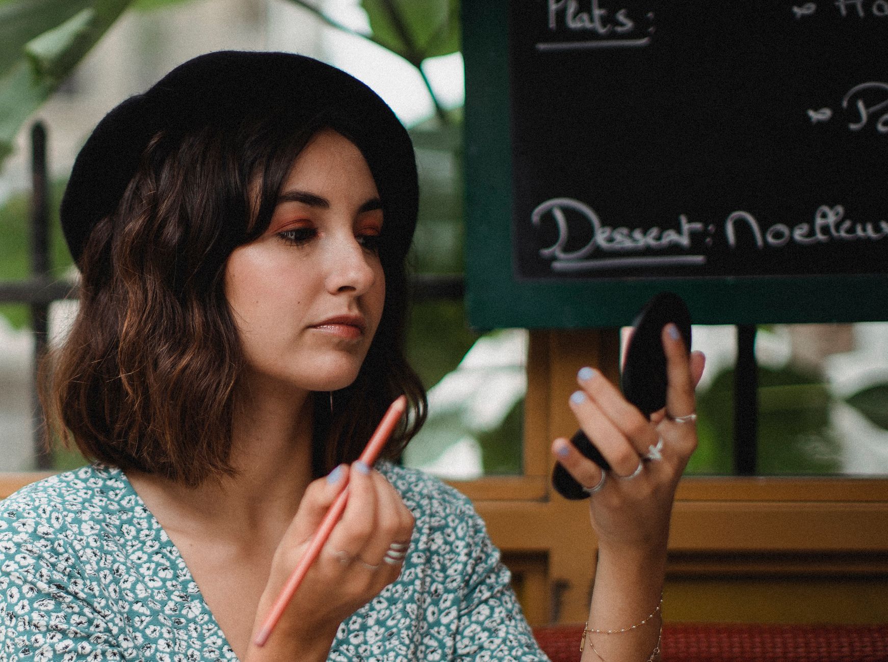 Young woman uses compact mirror for makeup touch-up in cozy café.