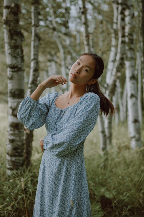 Dreamy Asian female wearing long dress standing among tall trees and grass in woods