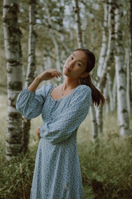 Dreamy Asian female wearing long dress standing among tall trees and grass in woods
