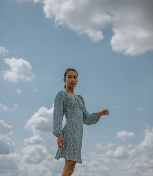 Low angle side view of serious Asian woman in elegant dress standing against cloudy sky
