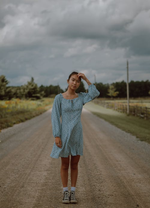 Full body of thoughtful Asian female in summer dress standing on empty road in rural area