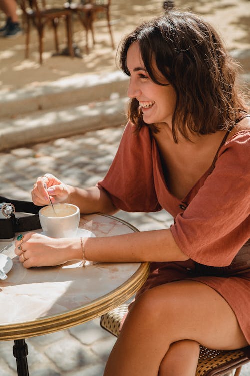 {{https://mixerdates.com/?ch=mixerblog}}Young woman smiling while stirring her coffee at an outdoor cafe table