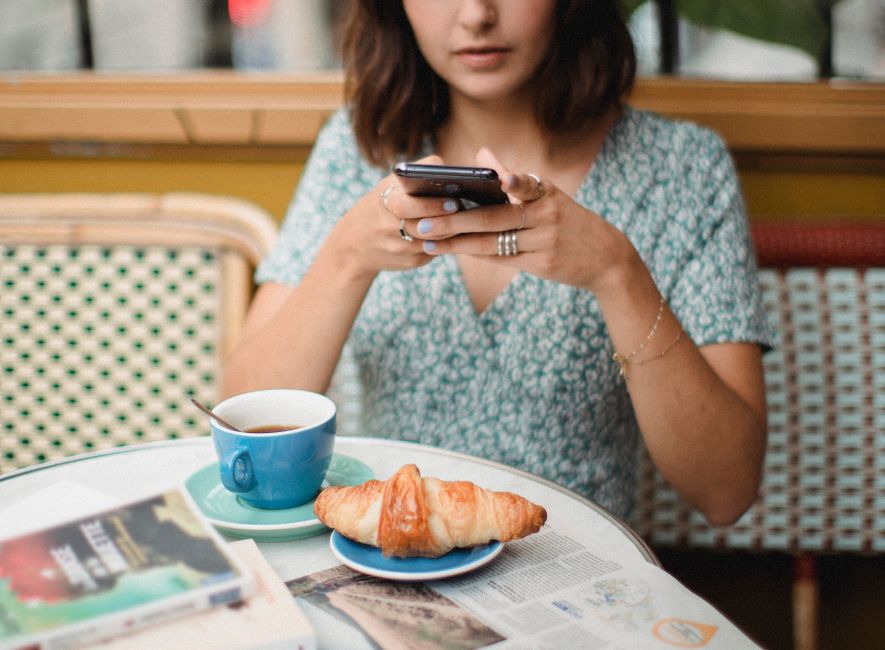 Woman taking photo of coffee and croissant in a cafe setting.