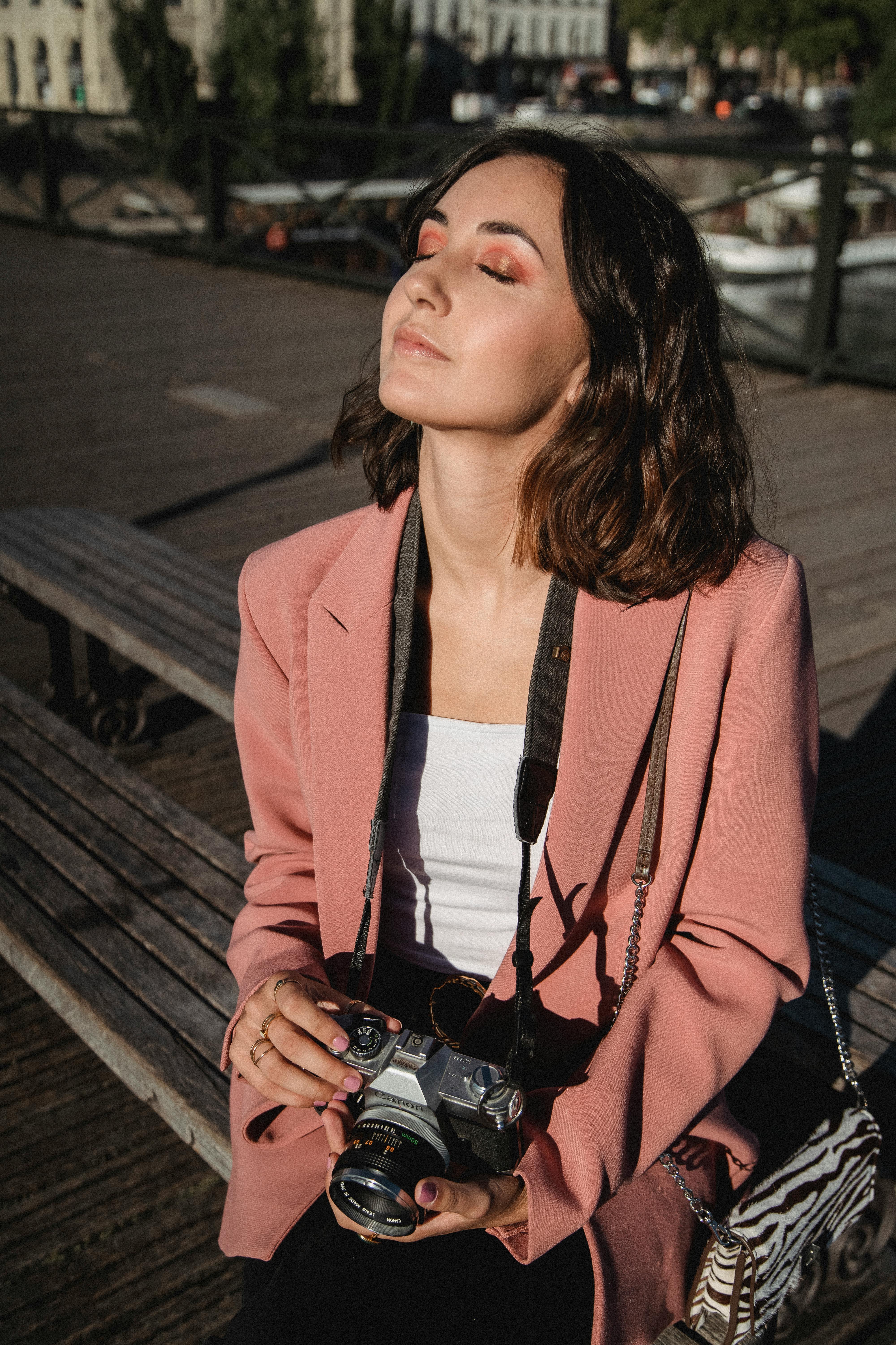Woman in pink blazer enjoying the sun on a wooden bench outdoors with a vintage camera.