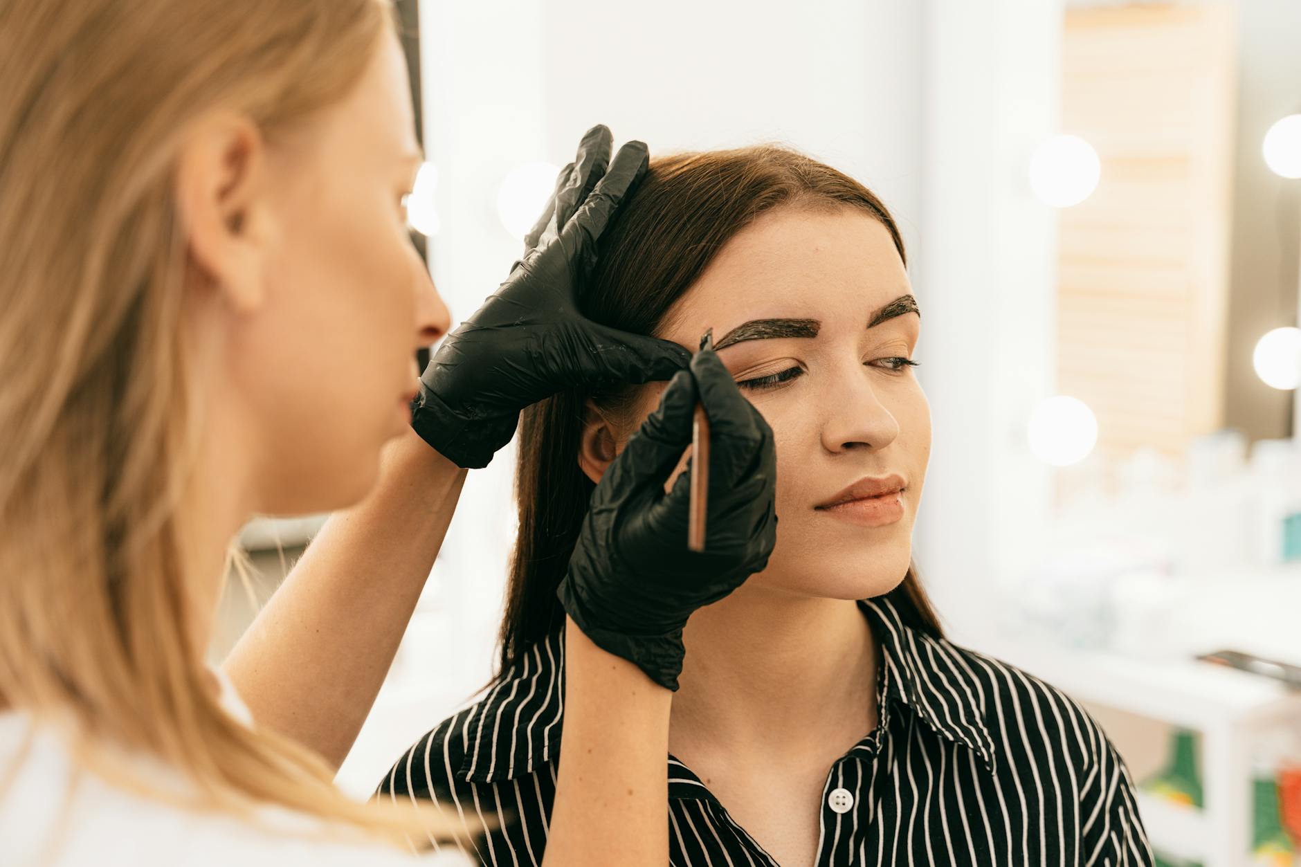 A professional makeup artist shapes a woman's eyebrows in a bright beauty studio.