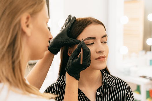 A professional makeup artist shapes a woman's eyebrows in a bright beauty studio.