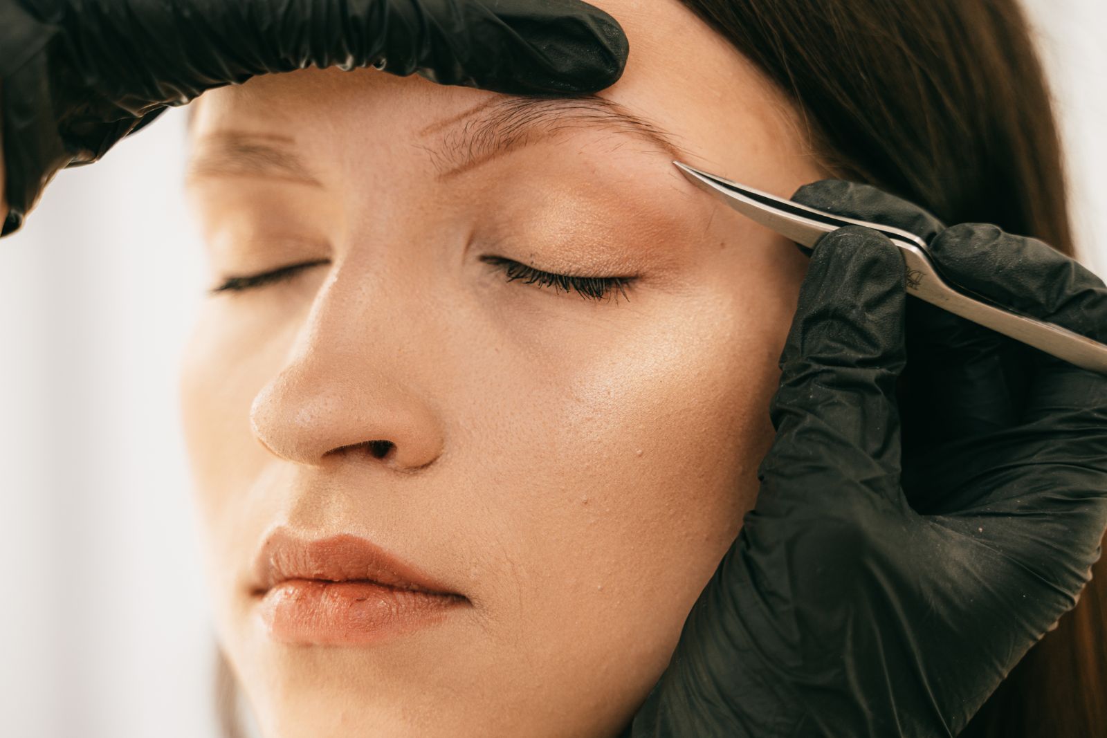 Close-up of a gloved beautician tidying a client's brow with tweezers