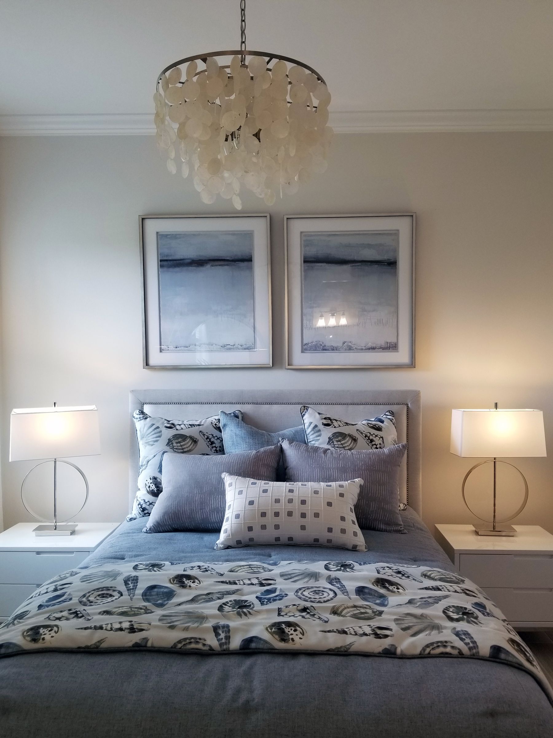 Blue-gray bedding and shell-patterned cushions beneath a gray headboard, two framed prints and a shell chandelier, with two lamps on white bedside tables