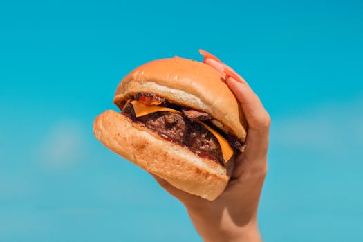 A close-up of a hand holding a delicious cheeseburger with a clear blue sky background.